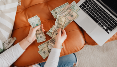Person counting cash beside a laptop on a cozy couch, representing simple ways to make extra money from home