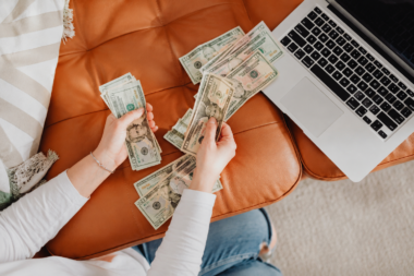 Person counting cash beside a laptop on a cozy couch, representing simple ways to make extra money from home