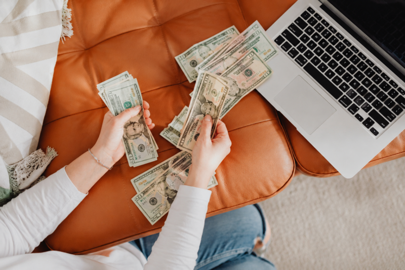 Person counting cash beside a laptop on a cozy couch, representing simple ways to make extra money from home